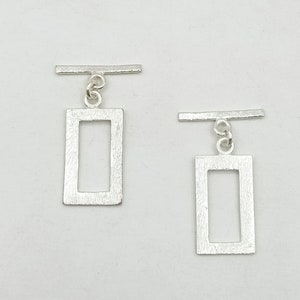 May include: A pair of silver earrings with a rectangular shape and a textured finish. Each earring features a small bar that hangs from the top of the rectangle.