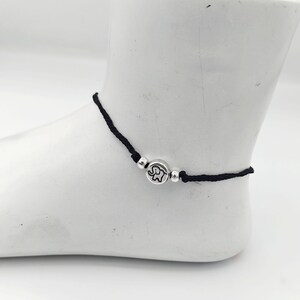May include: A black cord anklet featuring a central silver-toned circular charm with an engraved elephant design. The charm is flanked by two small, smooth silver-toned spherical beads, creating a simple and minimalist piece of jewelry.