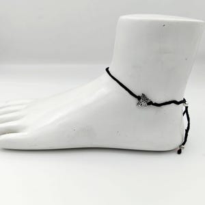 Op de afbeelding: Een enkelbandje van zwart koord met een kleine, gedetailleerde zilverkleurige vlinderbedel. Het enkelbandje wordt vastgemaakt met een eenvoudige knoop en is voorzien van kleine zilverkleurige kraaltjes aan de uiteinden van het verstelbare koord.