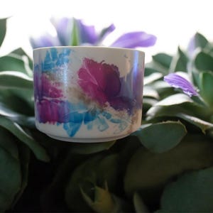 May include: A small, cylindrical ceramic cup with a floral design in shades of purple, blue, and brown. The cup is set against a backdrop of green succulent plants and purple flowers, creating a natural and artistic composition.