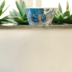 May include: A small, handmade ceramic bowl with a unique, abstract design. The bowl features a blend of blue, white, and gray colors, creating a marbled effect. The bowl is displayed in front of green succulent plants.