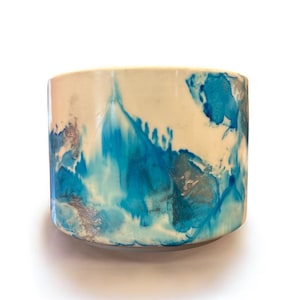 May include: A cylindrical ceramic planter with a cream-colored base and a vibrant blue and silver abstract design. The colors blend and drip down the sides, creating a unique, artistic look. The planter is suitable for indoor or outdoor use.
