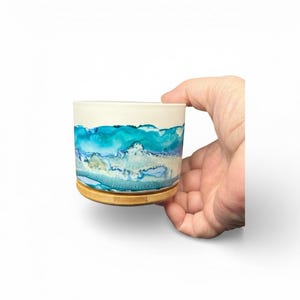 May include: A ceramic cup with a white top and a blue and turquoise wave design. The cup sits on a small, round, wooden coaster. The cup is held by a hand against a white background.