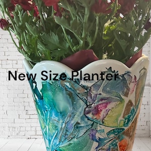 May include: A white ceramic planter with a scalloped rim, filled with red flowers and green foliage. The planter has a colorful abstract design in shades of blue, green, purple, and brown. Text on the planter reads "New Size Planter."