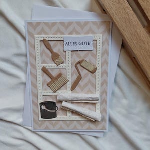 May include: A handmade greeting card with a beige and white chevron pattern background. The card features cut-out images of painting tools, including brushes, a paint roller, and a paint can. The text "ALLES GUTE" is displayed on a small white rectangle.
