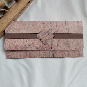 Puede incluir: Una cartera rectangular de papel texturizado marrón y beige, con una banda marrón oscuro y un acento cuadrado decorativo. La cartera está cerrada y parece estar hecha de papel hecho a mano. El fondo es una tela blanca.
