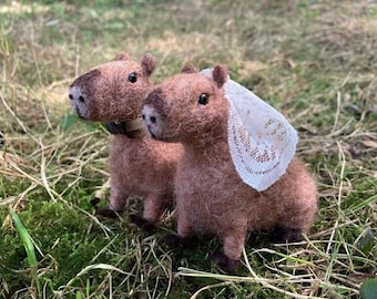 May include: Two brown felt capybara figurines, one wearing a white lace veil, standing on a bed of green moss.