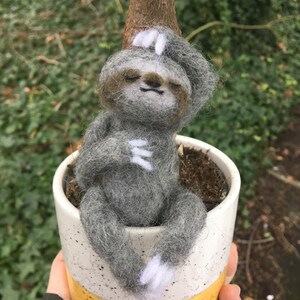 May include: A handmade felt sloth figurine in a small ceramic pot. The sloth is gray with white accents and appears to be relaxing. The pot is white with a yellow base and a speckled design. The background is blurred greenery.