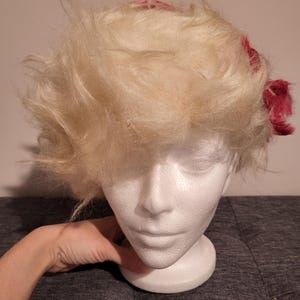 Puede incluir: Una peluca blanca con una diadema de patrón rojo y blanco. La peluca está peinada con un estilo desordenado y voluminoso.