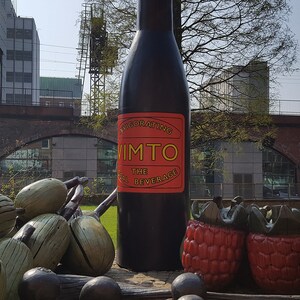 Puede incluir: Una gran botella negra con una etiqueta roja y amarilla que dice "Invigorating Vimto The Real Beverage". La botella est&aacute; rodeada de hierba verde y algunas esculturas de fruta grandes, de madera, rojas y verdes.