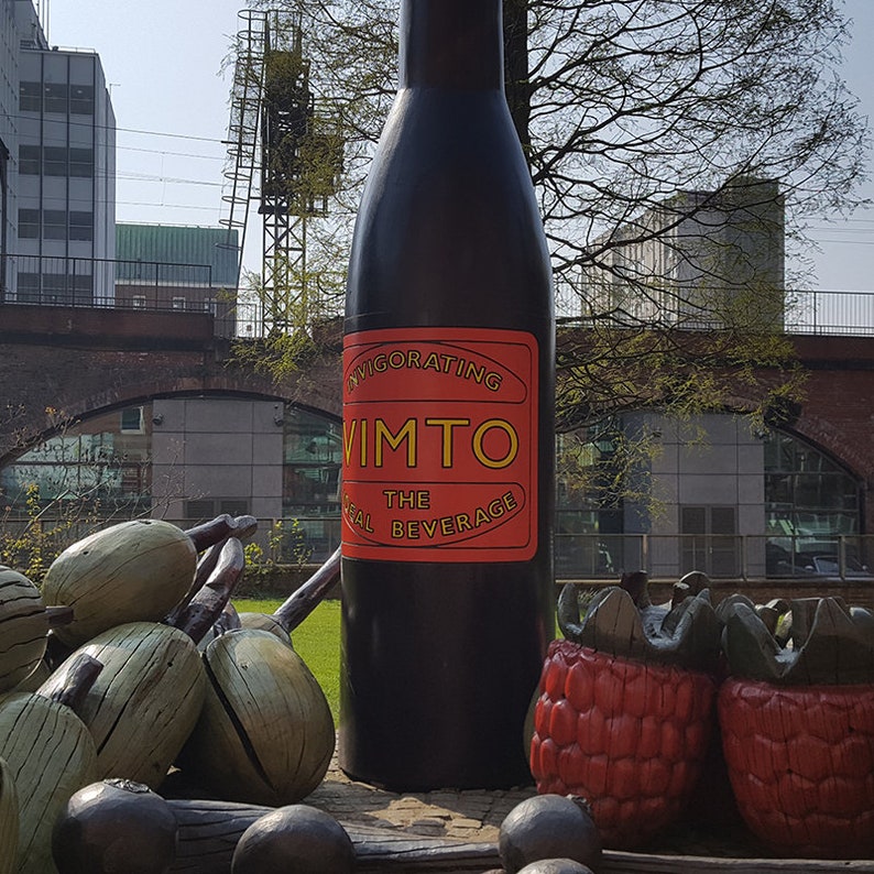 Puede incluir: Una gran botella negra con una etiqueta roja y amarilla que dice "Invigorating Vimto The Real Beverage". La botella est&aacute; rodeada de hierba verde y algunas esculturas de fruta grandes, de madera, rojas y verdes.