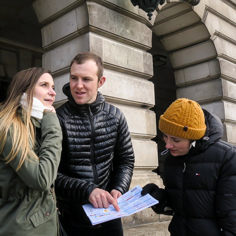 Puede incluir: Tres personas, dos mujeres y un hombre, est&aacute;n de pie frente a un edificio. El hombre sostiene un mapa y se&ntilde;ala algo en &eacute;l. La mujer de la izquierda lleva una chaqueta verde y una bufanda blanca. La mujer de la derecha lleva una chaqueta negra y una boina amarilla.