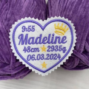 Op de afbeelding: Witte hartvormige viltpatch met paarse stiksels. De patch is geborduurd met een kroon, sterren en de tekst "9:55 Madeline 48cm 2935g 06.03.2024" in goud.