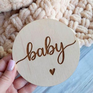 May include: A round wooden disc with the word "baby" engraved in a flowing script, accompanied by a small heart. The disc is light-colored and held against a soft, beige blanket.
