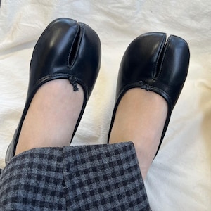 Op de afbeelding: Een paar zwarte leren split-teen ballerina's. De schoenen hebben een ronde neus met een split voor de grote teen en een klein decoratief detail aan de bovenkant. De ballerina's worden gedragen met een grijs-zwarte geruite broek.