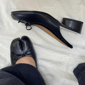 May include: Black leather split-toe shoes with a small bow detail. The shoes have a low, block heel and a unique design. The style is a modern take on a classic design. The shoes are shown on a white surface.