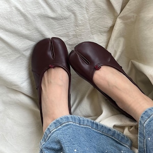 May include: Burgundy leather split-toe flats. The shoes have a low profile with a small red accent at the top. The flats are shown on a white fabric background, with the lower legs and feet of a person wearing blue jeans.