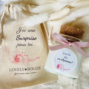 Peut inclure: Une petite bougie blanche dans un pot en verre avec un couvercle en liège. Le pot est attaché avec un ruban rose et a un autocollant rond avec le texte "Veux-tu etre ma Marraine?" et des motifs floraux roses. La bougie est dans un petit sac en toile blanche avec le texte "J'ai une Surprise pour toi... LOVELY BOUGIE fait main avec amour."