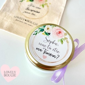 Peut inclure: Un pot de bougie blanc avec un couvercle doré et un motif floral. Le couvercle a un cœur rose et le texte "Sophie, veux-tu être ma Témoin?" dans un cercle doré.