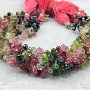 Puede incluir: Un collar de piedras preciosas multicolor con cuentas en forma de lágrima en tonos rosa, verde y negro. El collar está adornado con borlas rosas y detalles metálicos plateados.