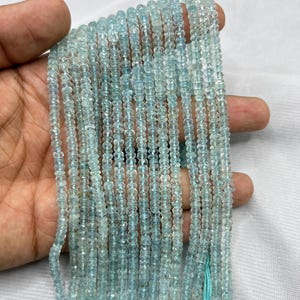 May include: A collection of light blue aquamarine gemstone beads. The beads are small, round, and faceted, creating a sparkling effect. The strands are held together with a light blue thread. The beads are a pale, translucent blue colour.