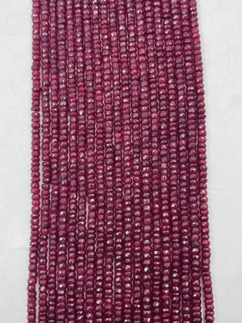May include: A strand of deep red ruby beads, each bead is faceted and has a slightly rounded shape. The beads are strung on a clear thread and are approximately 3mm in diameter.