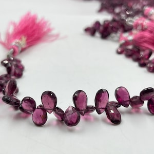 Könnte beinhalten: Eine Strähne aus violetten Edelsteinperlen, jede Perle hat die Form einer Träne. Die Perlen sind auf einem durchsichtigen Faden aufgefädelt und an jedem Ende mit rosa Quasten verziert.