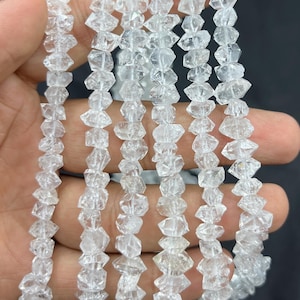 Op de afbeelding: Strengen heldere, gefacetteerde kwarts kralen. De kralen zijn aan elkaar geregen en vormen meerdere strengen. Het kwarts heeft een kristallijn uiterlijk en reflecteert licht. De kralen worden in een hand gehouden tegen een donkere achtergrond.
