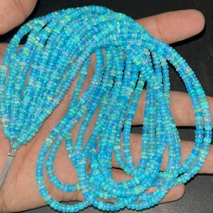 Pode incluir: Fios de contas de opala azul claro. As contas, pequenas e redondas, têm um brilho ligeiramente iridescente. Os fios são mantidos juntos em uma extremidade com um fio prateado.