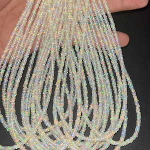 Op de afbeelding: Een close-up afbeelding van een streng witte opaal kralen, met iriserende kleurvlekken. De kralen zijn klein en rond, en ze zijn geregen op een doorzichtige draad.