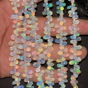Peut inclure: Plusieurs brins de perles d'opale iridescentes. Les perles en forme de goutte d'eau sont enfilées ensemble, affichant une gamme de couleurs comprenant le blanc, le bleu, le vert et l'orange. Les pierres précieuses sont tenues dans une main.