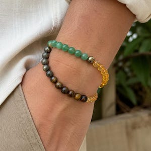 Puede incluir: Una pulsera de cuentas con una secuencia de cuentas redondas verdes, gris metálico, marrones y amarillas translúcidas. La pulsera está construida con secciones alternas de piedras lisas y pulidas en variados tonos tierra y colores vivos.