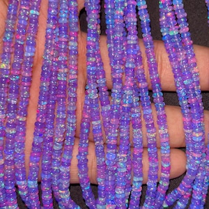 Könnte beinhalten: Nahaufnahme von mehreren Strängen lila Opalperlen. Die kleinen, runden Perlen haben einen schillernden Glanz mit rosa, blauen und grünen Blitzen. Die Perlen sind aufgefädelt und bilden eine strukturierte und farbenfrohe Darstellung.