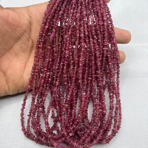 Puede incluir: Múltiples hebras de pequeñas piedras preciosas irregulares de color rosa intenso. Las cuentas están ensartadas, creando múltiples hebras. Las piedras preciosas tienen una textura rugosa y natural y un color rico y saturado. El fondo es una superficie blanca lisa.
