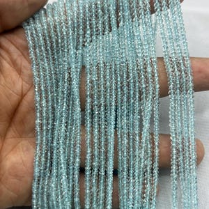 Könnte beinhalten: Mehrere Stränge kleiner, hellblauer Edelsteinperlen. Die Perlen sind aufgereiht und bilden vertikale Linien. Die Perlen sind ein durchscheinendes, helles Blau und scheinen facettiert zu sein. Die Stränge werden unten von einer türkisfarbenen Schnur zusammengehalten.