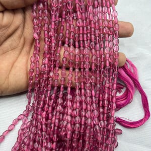 Può includere: Una collana di perline ovali rosa di pietra preziosa. Le perline sono traslucide e hanno un leggero luccichio. Le perline sono infilate su un filo trasparente.