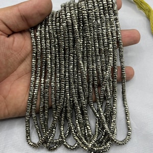 May include: Strands of small, metallic gray beads. The beads are rectangular and strung together, forming multiple strands. A yellow-green string is visible at the top. The beads are held in a hand against a white background.