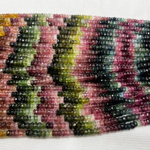 Puede incluir: Una colección de cuentas de turmalina facetadas en forma de rondel, dispuestas en múltiples hilos paralelos. Las cuentas muestran un espectro vibrante de colores, incluyendo tonos de rosa, verde, amarillo, marrón y azul profundo, con un acabado pulido.