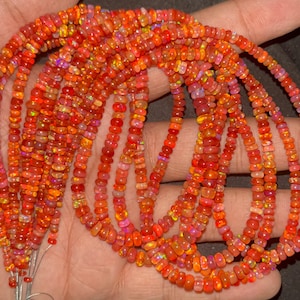 May include: A strand of orange opal beads, with a mix of light and dark orange hues, is displayed on a hand. The beads are small and round, and they have a smooth, polished finish.