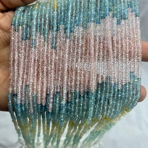 Könnte beinhalten: Stränge aus kleinen, facettierten Perlen in Rosa-, Blau- und Gelbtönen. Die Perlen sind aufgefädelt und erzeugen einen strukturierten, geschichteten Effekt. Die Perlen werden in einer Hand gehalten, wodurch die Farbvariationen und die zarte Natur des Materials hervorgehoben werden.