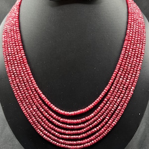 May include: A multi-strand necklace made of small, faceted red beads. The necklace is displayed on a black mannequin, highlighting the layered design. The beads are uniform in size and shape, creating a consistent appearance. The necklace is a deep red colour.