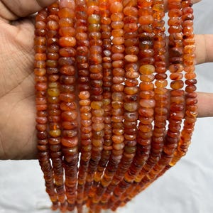 May include: Multiple strands of orange-red gemstone beads. The small, faceted beads are strung together, showing colour variations from light orange to deep red. The beads are held in a hand against a white background.