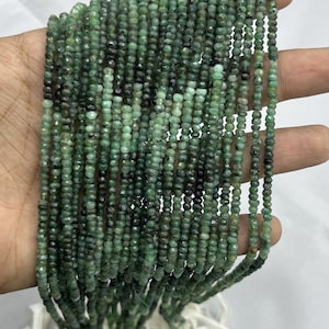 May include: Strands of small, faceted emerald beads in varying shades of green and dark green. The beads are strung together, with white string visible at the bottom. The beads are held in a hand against a white background.
