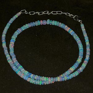 May include: A delicate necklace composed of small, iridescent opal beads. The beads display a range of colors, including blue, green, and pink, strung together with a silver chain and clasp. The necklace is displayed against a dark background.