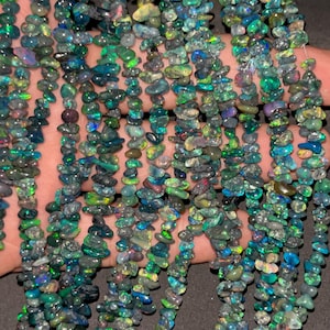 Può includere: Primo piano di fili di schegge di opale iridescenti. Le schegge sono prevalentemente verdi, blu e nere, con lampi di altri colori. Le schegge sono infilate insieme, creando un effetto scintillante. Le perle sono piccole e di forma irregolare.