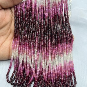May include: Strands of small, faceted beads in a gradient of colors from deep burgundy to light pink and white. The beads are strung together, creating a textured and colorful display. The beads are likely gemstones, perfect for jewelry making.