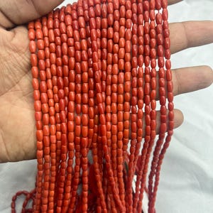 Op de afbeelding: Een streng levendige rode koraal kralen. De kralen zijn klein, cilindrisch en aan elkaar geregen, waardoor een vloeiend, gestructureerd uiterlijk ontstaat. Het koraal heeft een gepolijste, glanzende afwerking, die licht reflecteert. De kralen worden in een hand gehouden, waarbij hun kleur en vorm worden getoond.