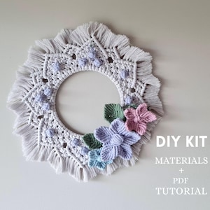 May include: DIY kit for a macrame wreath with flowers. The wreath is white with a fringe border and features a floral design with pink, purple, and blue flowers. The kit includes materials and a PDF tutorial.