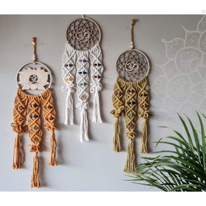 Può includere: Tre acchiappasogni in macramè con cornici in legno e accenti di pietre preziose colorate sono appesi a una parete grigia. Gli acchiappasogni presentano intricate nodi, nappe e simboli come il simbolo Om e un fiore di loto.