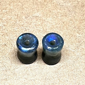 May include: A pair of iridescent blue and black ear gauges. The gauges are made of a smooth, polished material and have a slightly flared shape.